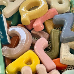 May include: A collection of ceramic alphabet letters in various colors and finishes, including glossy yellow, pink, blue, and speckled white, packed inside a light green plastic crate with a grid pattern base.
