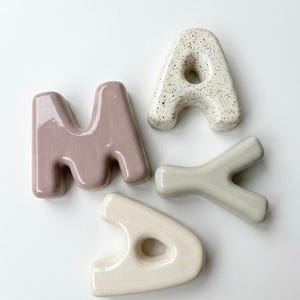 May include: Four ceramic letters spelling MAYA. The letter M is mauve, the letter A is white with black speckles, the letter Y is sage green, and the second letter A is cream. Each letter has a smooth, glossy finish and rounded, bubble-like edges.