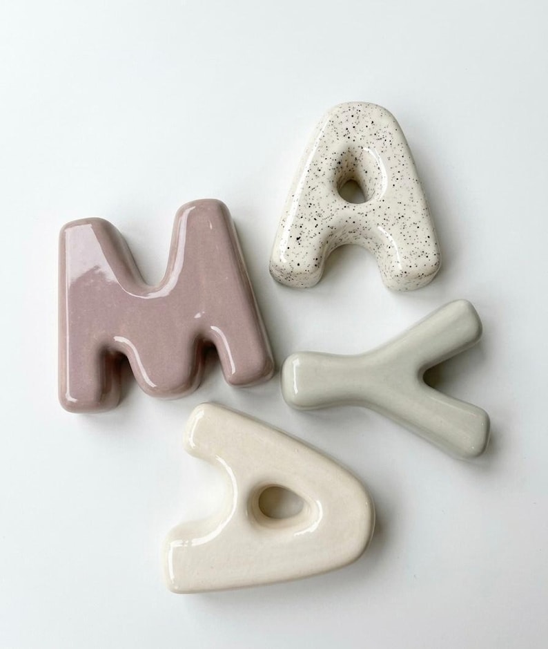 May include: Four ceramic letters spelling MAYA. The letter M is mauve, the letter A is white with black speckles, the letter Y is sage green, and the second letter A is cream. Each letter has a smooth, glossy finish and rounded, bubble-like edges.