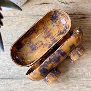 May include: Open ceramic serving dish with a unique design. The dish is a warm orange color with dark brown accents, creating a textured, mottled appearance. The dish is oval-shaped with a lid, and sits on two rounded feet.