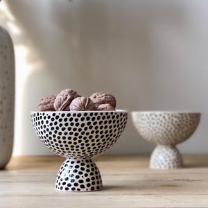 Może przedstawiać: Dwie ceramiczne miski z czarno-białym wzorem w kropki. Jedna miska jest wypełniona orzechami włoskimi. Miski mają podstawę. Tło obejmuje biały wazon i drewnianą powierzchnię.