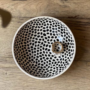 Op de afbeelding: Witte keramische kom met een zwart stippenpatroon en een gouden ring in het midden. De kom staat op een houten oppervlak.