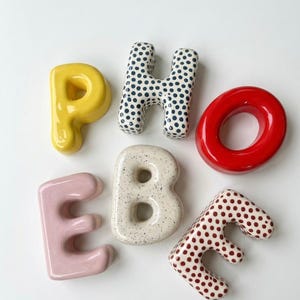 Op de afbeelding: Een verzameling keramische letters die "PHOEBE" spellen. De letters zijn in verschillende kleuren en patronen, waaronder geel, stippen, rood, roze, gespikkeld wit en bruine stippen. De letters zijn gerangschikt op een witte ondergrond.