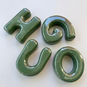 Pode incluir: Quatro letras individuais de cerâmica com um acabamento verde-oliva brilhante que formam a palavra HUGO. As letras estão dispostas em grupo, apresentando bordas arredondadas e uma superfície lisa e reflexiva que capta a luz nas formas.