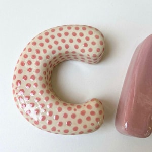 May include: A ceramic letter C with a glossy cream-colored finish and a pattern of small, raised pink polka dots, positioned next to a portion of a solid, smooth, glossy mauve-colored ceramic letter, both featuring rounded edges and a thick profile.