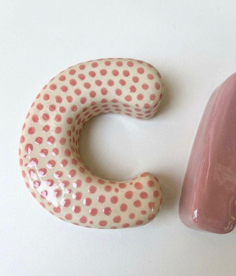 May include: A ceramic letter C with a glossy cream-colored finish and a pattern of small, raised pink polka dots, positioned next to a portion of a solid, smooth, glossy mauve-colored ceramic letter, both featuring rounded edges and a thick profile.