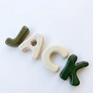 May include: Four individual ceramic letters spelling the name JACK. The letter J is olive green, the A is cream with black speckles, the C is solid cream, and the K is dark forest green. Each letter has a smooth, glossy, rounded finish.