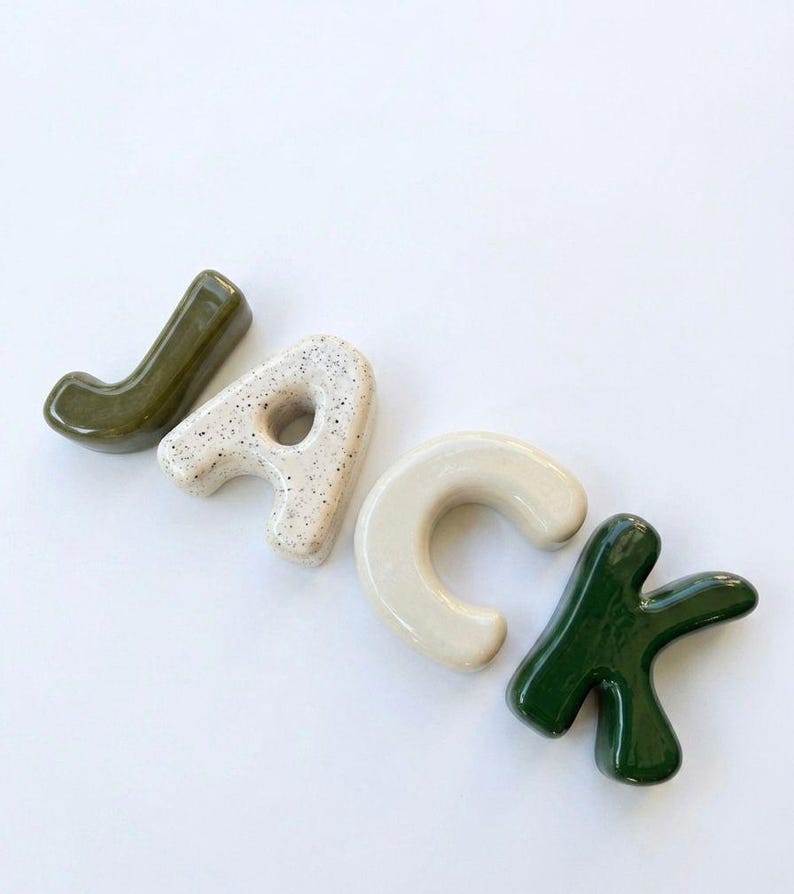 May include: Four individual ceramic letters spelling the name JACK. The letter J is olive green, the A is cream with black speckles, the C is solid cream, and the K is dark forest green. Each letter has a smooth, glossy, rounded finish.