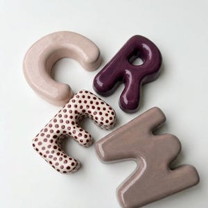 May include: Four ceramic letters spelling the word "CREH" in a rounded, bubbly font. The letter C is light pink, the R is deep purple, the E is white with brown polka dots, and the H is a muted mauve. Each letter has a smooth, glossy finish.