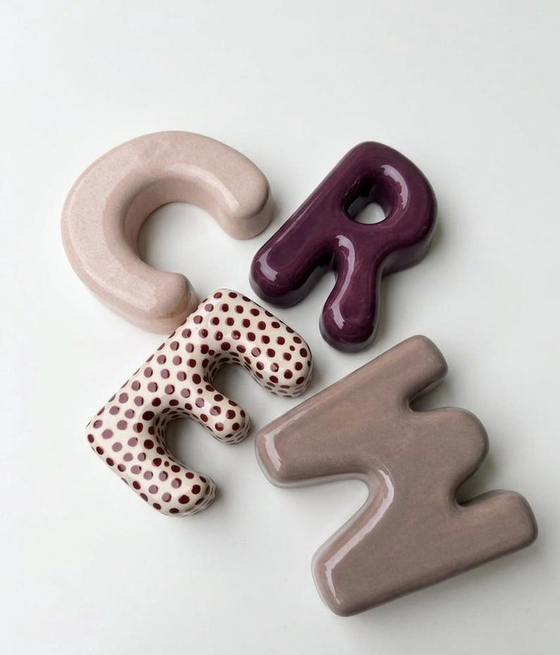May include: Four ceramic letters spelling the word "CREH" in a rounded, bubbly font. The letter C is light pink, the R is deep purple, the E is white with brown polka dots, and the H is a muted mauve. Each letter has a smooth, glossy finish.