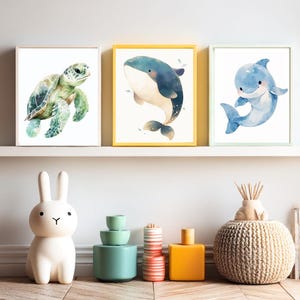 以下が含まれることがあります： 緑色のウミガメ、青いクジラ、青いイルカの3つの額入り水彩画。白い棚に飾られています。クジラのプリントは黄色のフレーム、イルカのプリントは薄緑色のフレーム、カメのプリントは白いフレームです。
