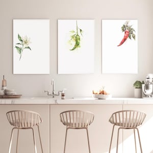 以下が含まれることがあります： 3つの額入り水彩画プリント。白い花、緑色の唐辛子、赤い唐辛子の植物画が描かれています。プリントは、バーのスツールのあるキッチンカウンターの上に表示されています。