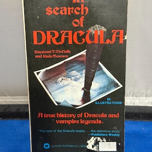 May include: A paperback book titled In Search of Dracula by Raymond T. McNally and Radu Florescu. The black cover features red text, a central illustration of a wooden stake piercing a figure, and the subtitle A true history of Dracula.