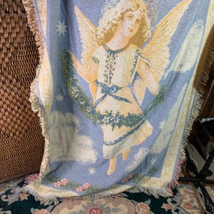 May include: A woven throw blanket featuring a celestial angel design. The angel has golden wings and a halo, set against a light blue backdrop with clouds and stars. The blanket has a fringed edge and a floral border.