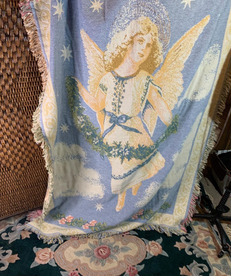 May include: A woven throw blanket featuring a celestial angel design. The angel has golden wings and a halo, set against a light blue backdrop with clouds and stars. The blanket has a fringed edge and a floral border.