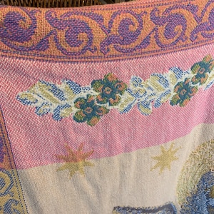 May include: Close-up of a woven textile with a detailed pattern. The design features a pink and cream background with floral and star motifs. The border has a repeating pattern in purple and orange.