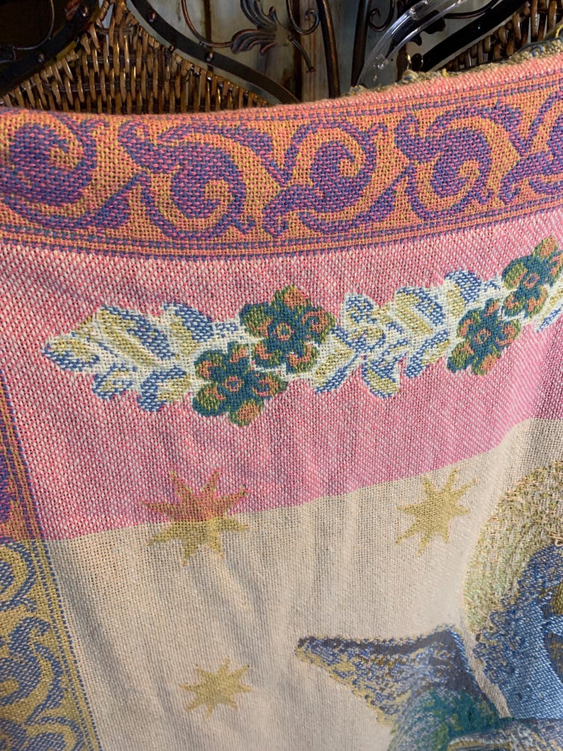 May include: Close-up of a woven textile with a detailed pattern. The design features a pink and cream background with floral and star motifs. The border has a repeating pattern in purple and orange.