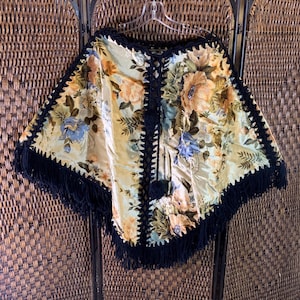 May include: A vintage-style poncho with a floral pattern in shades of yellow, orange, and blue. The edges are trimmed with black crochet and fringe. The poncho is hanging on a clear hanger.