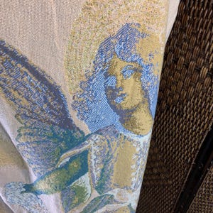 May include: A woven textile featuring a detailed angel design. The angel has a halo and wings, rendered in shades of blue, gold, and green against a beige background. The textile is folded, revealing a glimpse of pink and white stripes.