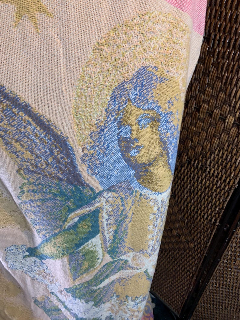 May include: A woven textile featuring a detailed angel design. The angel has a halo and wings, rendered in shades of blue, gold, and green against a beige background. The textile is folded, revealing a glimpse of pink and white stripes.