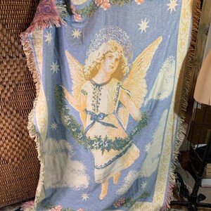 May include: A light blue woven throw blanket featuring an angel with golden wings, a halo, and a white dress. The blanket has a floral border and star accents. The edges are fringed with pink and yellow.