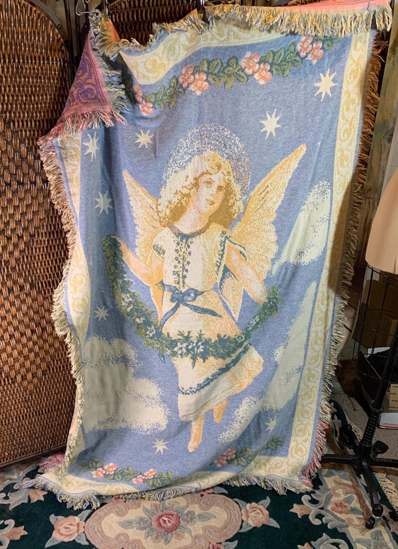 May include: A light blue woven throw blanket featuring an angel with golden wings, a halo, and a white dress. The blanket has a floral border and star accents. The edges are fringed with pink and yellow.