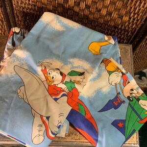 May include: Light blue vintage bed sheet featuring cartoon characters in flight. The sheet has a repeating pattern of cartoon characters, clouds, and a rocket ship. The characters are in red, yellow, and green. The sheet is folded.