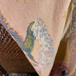 May include: A woven tapestry blanket with a peach background and a detailed landscape design. The design includes trees, stars, and decorative borders in shades of blue, green, and yellow. The blanket has a fringed edge.