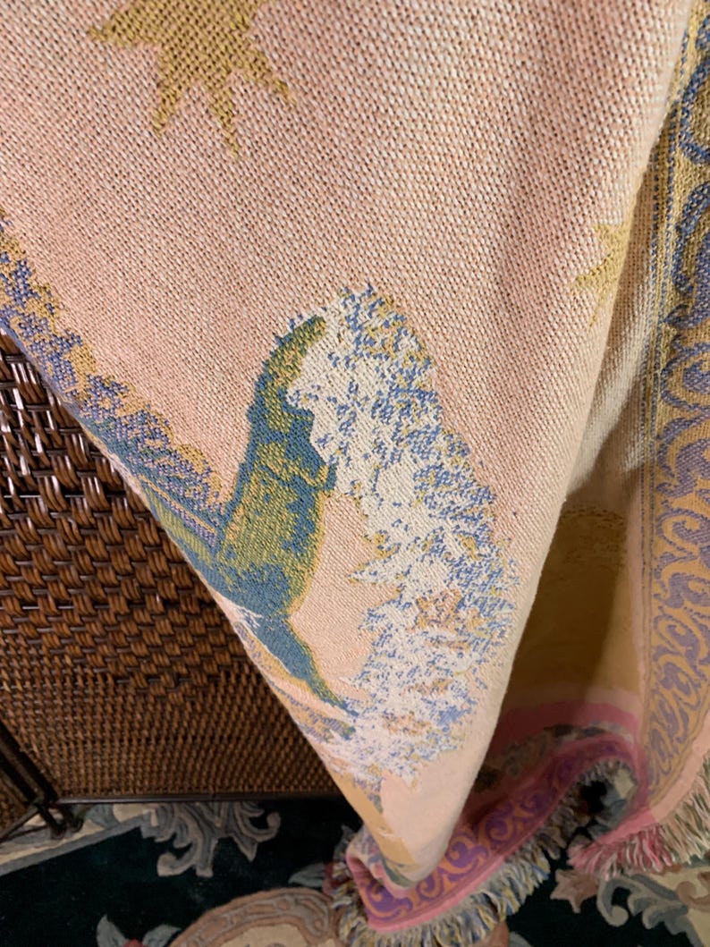 May include: A woven tapestry blanket with a peach background and a detailed landscape design. The design includes trees, stars, and decorative borders in shades of blue, green, and yellow. The blanket has a fringed edge.