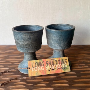 May include: Two blue ceramic goblets with a textured finish, each with a wide bowl and a pedestal base. A tag with the text "LONG SHADOWS Vintage" is attached to the goblets. The goblets are displayed on a wooden surface.