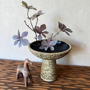 May include: A ceramic pedestal bowl with a black interior holds branches with purple-tinged leaves. A small, brown, carved horse figurine stands nearby. The bowl has a speckled, beige exterior and a wide, shallow design.