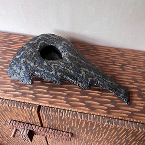 May include: A sculptural ceramic object with a dark, textured, and uneven surface featuring deep black and blue tones. The piece has an elongated, tapered shape with a prominent circular opening near the wider end, showcasing a rough, organic finish.