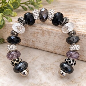 May include: A cuff bracelet featuring a variety of beads including smooth black, faceted dark gray, clear crackle glass, purple crackle glass, silver-toned metal, and pave-set clear crystal beads arranged in a symmetrical pattern.