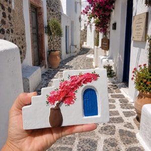 May include: A rectangular white ceramic plaque with a stepped top edge, featuring a hand-painted scene of a brown potted plant with vibrant pink flowers next to a small, arched blue door, set against a white wall background.