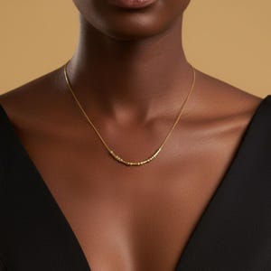 Könnte beinhalten: Eine zarte, goldfarbene Halskette mit einer Reihe kleiner, gleichmäßig verteilter Perlen. Die Halskette wird vor einem dunklen Hintergrund getragen, was einen auffälligen Kontrast erzeugt. Die Halskette ist ein schlichtes, aber elegantes Schmuckstück.