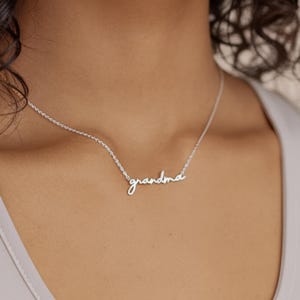 May include: A silver-toned necklace featuring a delicate chain and a central pendant in cursive script that reads "grandma". The metal has a polished finish, and the pendant is integrated directly into the chain, resting against the neck.