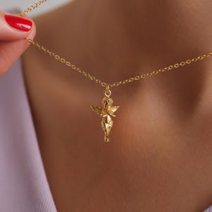 Puede incluir: Un collar dorado con un colgante de querub&iacute;n. El querub&iacute;n, con alas detalladas, cuelga de una fina cadena. El collar se sostiene, destacando el dise&ntilde;o y el color. El fondo est&aacute; borroso, atrayendo la atenci&oacute;n hacia la joya. La cadena y el colgante son dorados.