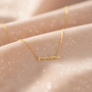 May include: A gold-toned necklace featuring a delicate chain and a central pendant formed by the word "grandma" written in a cursive, lowercase script font. The metal has a polished finish that reflects light, creating a bright, metallic appearance.