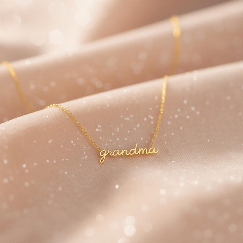 May include: A gold-toned necklace featuring a delicate chain and a central pendant formed by the word "grandma" written in a cursive, lowercase script font. The metal has a polished finish that reflects light, creating a bright, metallic appearance.
