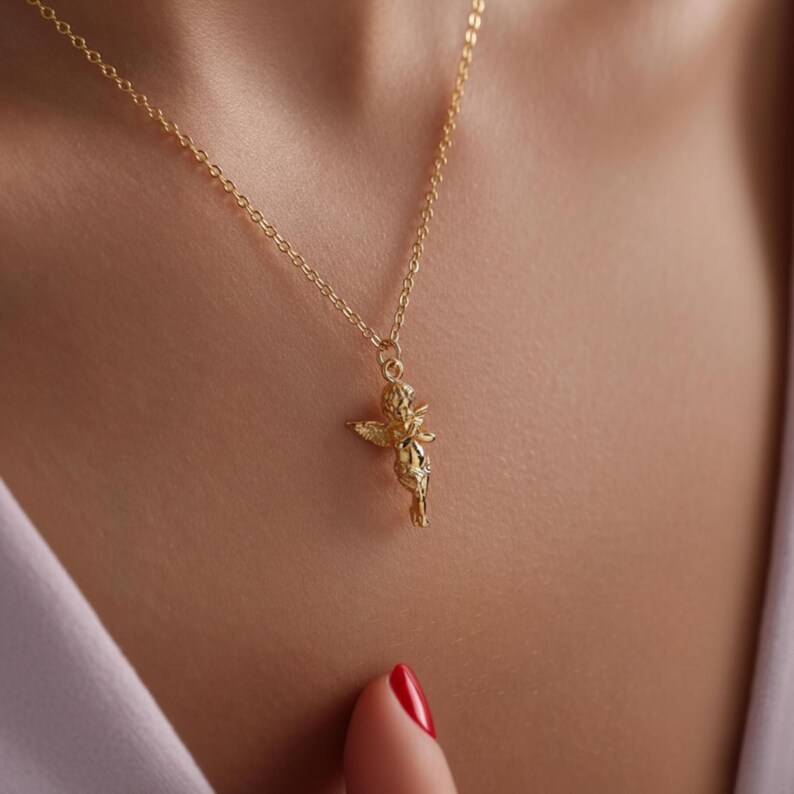 Puede incluir: Un collar dorado con un peque&ntilde;o colgante de querub&iacute;n. El querub&iacute;n tiene alas extendidas y detalles finos. La delicada cadena cuelga contra la piel de una persona, creando un aspecto simple pero elegante.