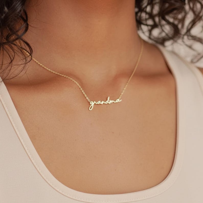 May include: A gold-toned necklace featuring a delicate chain and a central pendant in cursive script that reads "grandma". The metal has a polished finish, and the pendant is integrated directly into the chain, resting against the collarbone area.
