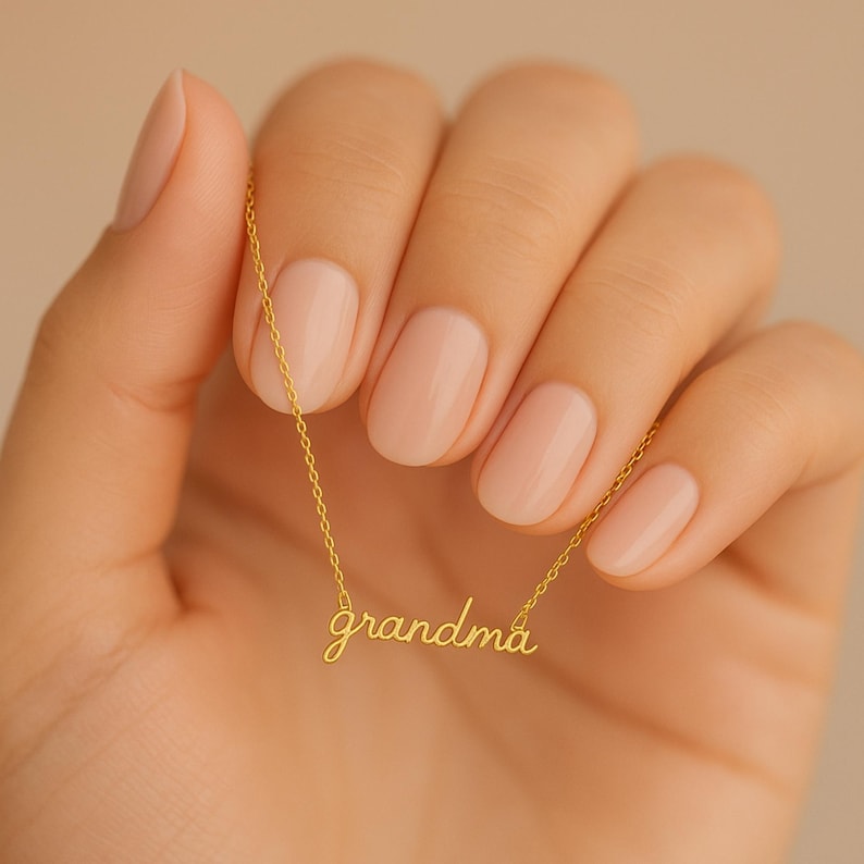 May include: A gold-toned necklace featuring a delicate chain and a central pendant in cursive script that spells out the word grandma. The metal has a polished finish, and the pendant is integrated directly into the thin, lightweight chain links.