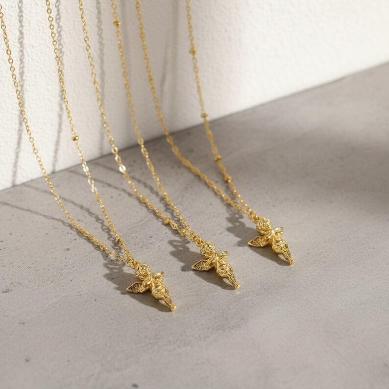 Puede incluir: Varias cadenas doradas con colgantes de &aacute;ngeles. Las cadenas son delicadas, con peque&ntilde;os detalles de cuentas. Cada colgante de &aacute;ngel est&aacute; detallado con alas y vestiduras fluidas. Se exhiben sobre una superficie gris claro.