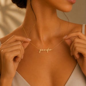 May include: A gold-toned metal necklace featuring a delicate chain and a central nameplate pendant displaying the name Jennifer in a cursive, flowing script font. The necklace rests against the collarbone, showcasing a polished, reflective finish.