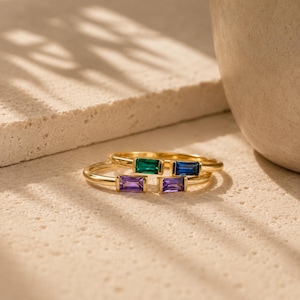 Könnte beinhalten: Zwei Goldringe mit im Baguetteschliff geschliffenen Edelsteinen. Ein Ring zeigt einen grünen und einen blauen Stein, während der andere Ring zwei violette Steine aufweist. Die Ringe haben ein dünnes, poliertes Goldband mit horizontal gefassten Steinen.