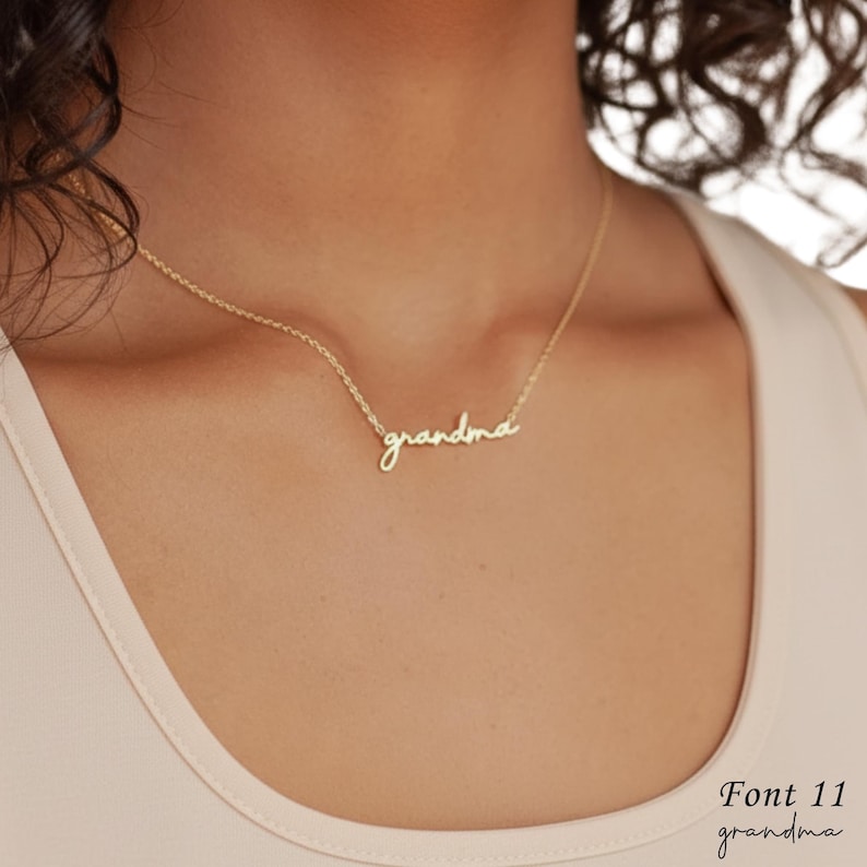 May include: A gold-toned necklace featuring a delicate chain and a cursive script pendant that spells out the word grandma. The pendant is centered on the chain, which rests against the collarbone area, showcasing a simple and elegant design.