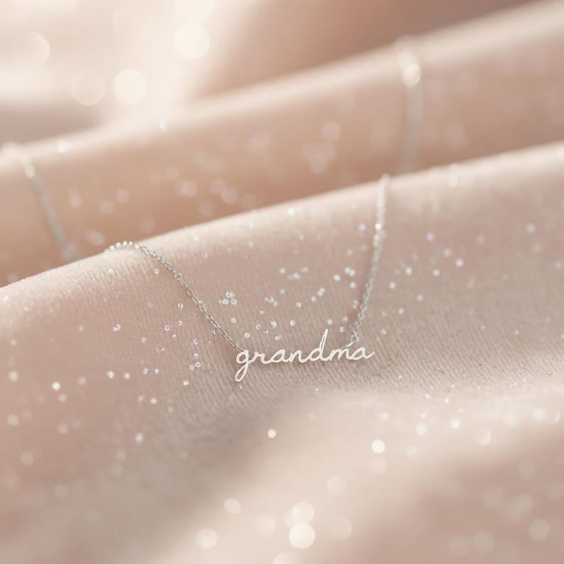 May include: A silver-toned metal necklace featuring a delicate chain and a script-style pendant that spells out the word grandma in lowercase cursive letters. The piece is crafted with a thin, polished metal finish and a simple, minimalist link chain.
