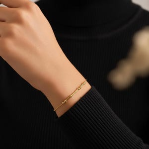 May include: A delicate gold bracelet featuring a series of small, round beads and cylindrical bars. The bracelet is worn on a wrist, with a black long-sleeved top in the background. The jewelry is simple and elegant.
