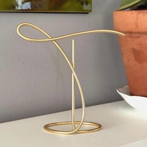 Op de afbeelding: Een goudkleurige metalen decoratieve standaard met een lusvormig ontwerp. De standaard heeft een cirkelvormige basis en een verticale steun met een gebogen bovenkant. Ontworpen om sieraden of kleine items vast te houden.