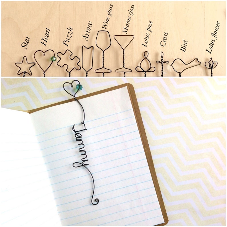 Lying on an opened book, a customizable bookmark made in black wire is shaped into a persons name in a decorative font.  It has a heart outline with a bead on it at the top, the heart extends above the page and the name runs vertical down the page.